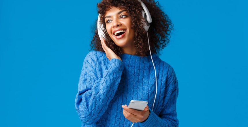 Charismatic modern young attractive african-american girl with afro haircut, listening music in headphones looking out window snowy weather, holding smartphone, smiling and singing.